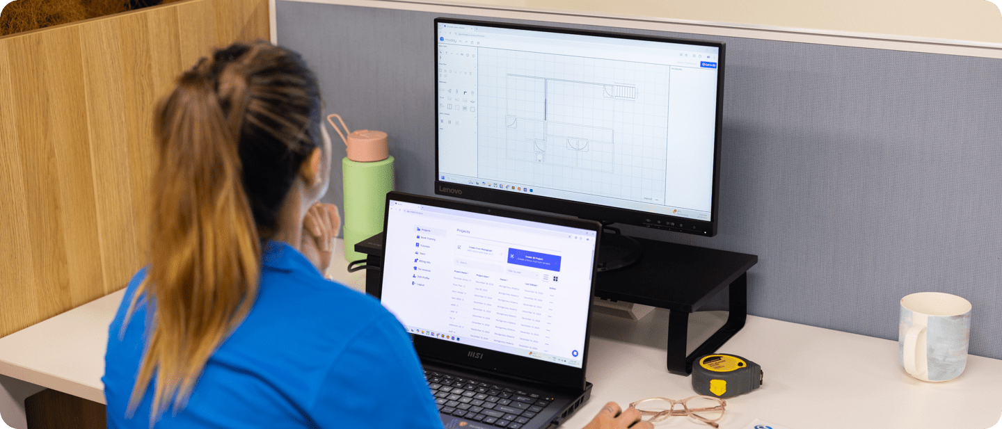 An occupational therapist with long brown hair tied in a ponytail, wearing a blue shirt, is seated at a white desk. They are working on a laptop showing a project management tool while viewing a monitor displaying "Moddy," a drawing software for occupational therapists. The desk contains a green water bottle with a pink lid, a measuring tape, glasses, and a coffee mug. The background features a wooden partition and a grey desk divider.