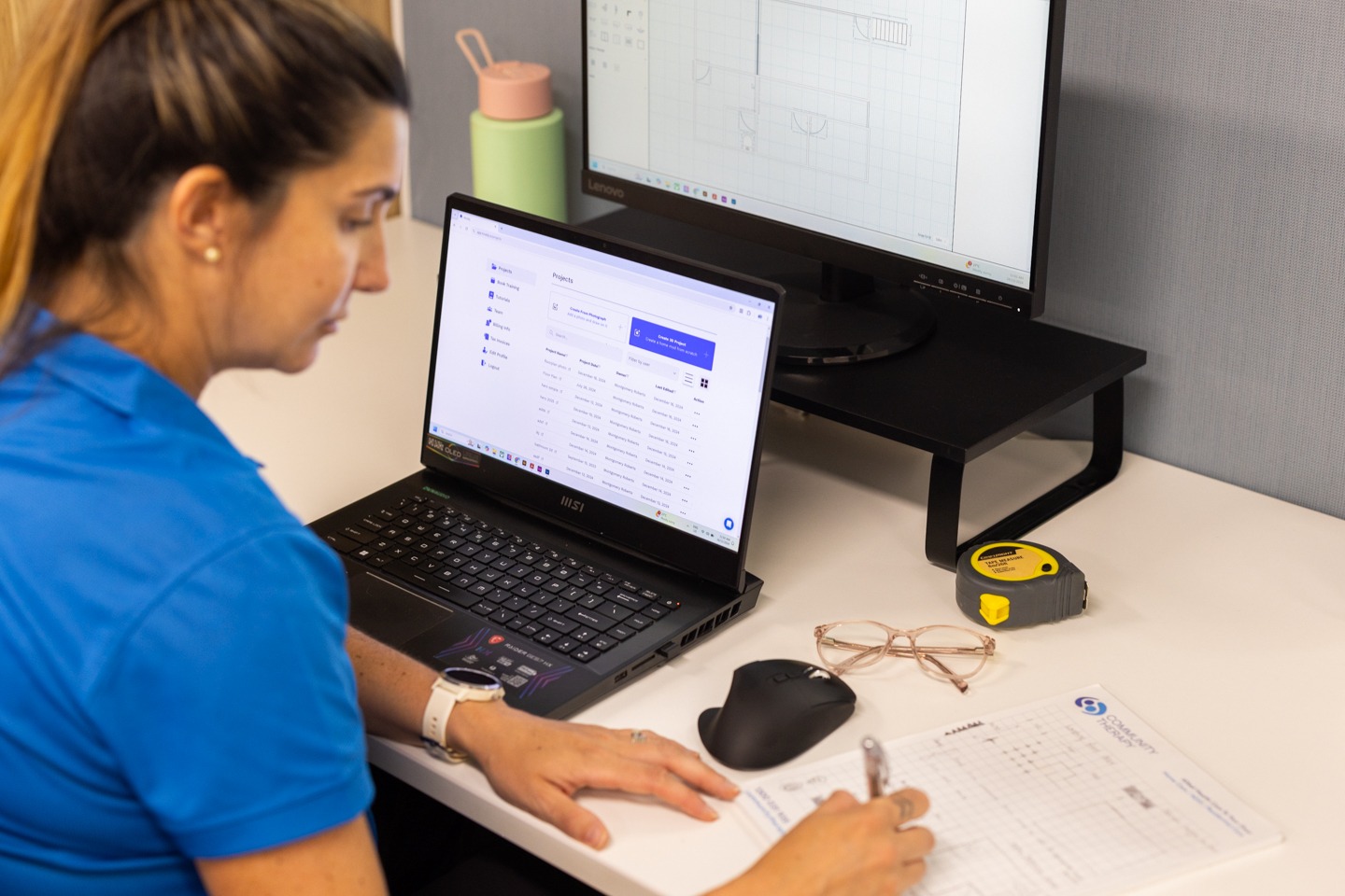 A woman with brown hair tied in a ponytail wearing a blue shirt is seated at a white desk, working on a project. They are writing on a paper document with a pen while looking at a laptop screen displaying Moddy - drawing software for Occupational Therapists. A black computer monitor on a stand shows a technical drawing. Nearby on the desk are a black computer mouse, a pair of glasses, a yellow measuring tape, and a green water bottle with a pink lid.