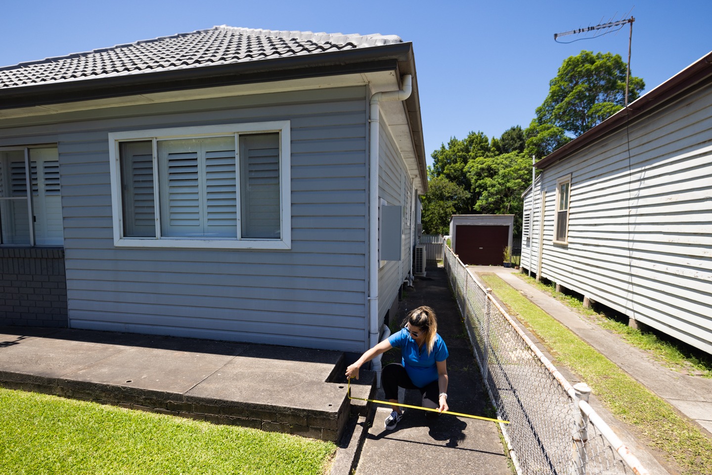 A woman in a blue shirt crouches down outside a single-story house with a grey facade, measuring a small step with a yellow measuring tape. A narrow pathway runs alongside the house, bordered by a chain-link fence. Another house is visible in the background, along with green grass and trees under a clear blue sky.