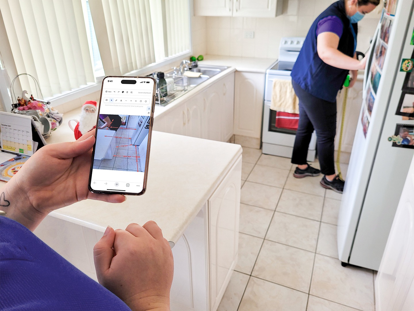 Person holding a phone displaying kitchen measurements while another person measures the fridge space